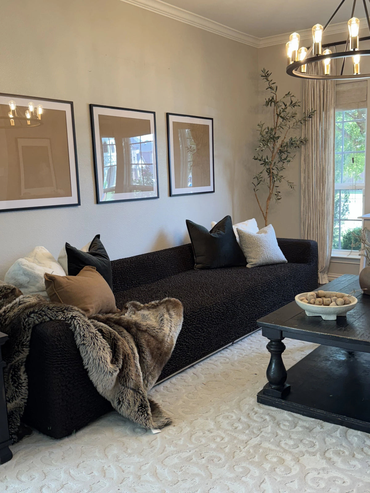 A dark textured boucle sofa in a styled living room, decorated with black, white, and brown throw pillows and a brown faux fur throw blanket. A black coffee table and a white patterned rug are in front of the sofa, with three framed art prints on the beige wall behind it | Influencers Welcome