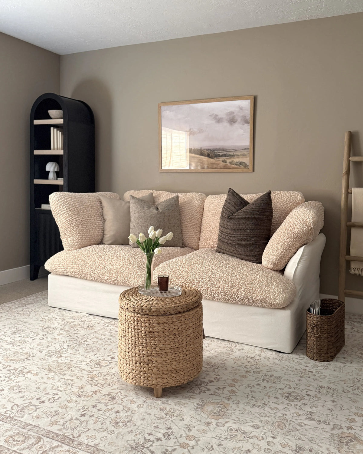 A cozy living room scene featuring a cream bouclé loveseat with textured throw pillows, a round woven ottoman table, a patterned area rug, and neutral-toned walls | Influencers Welcome