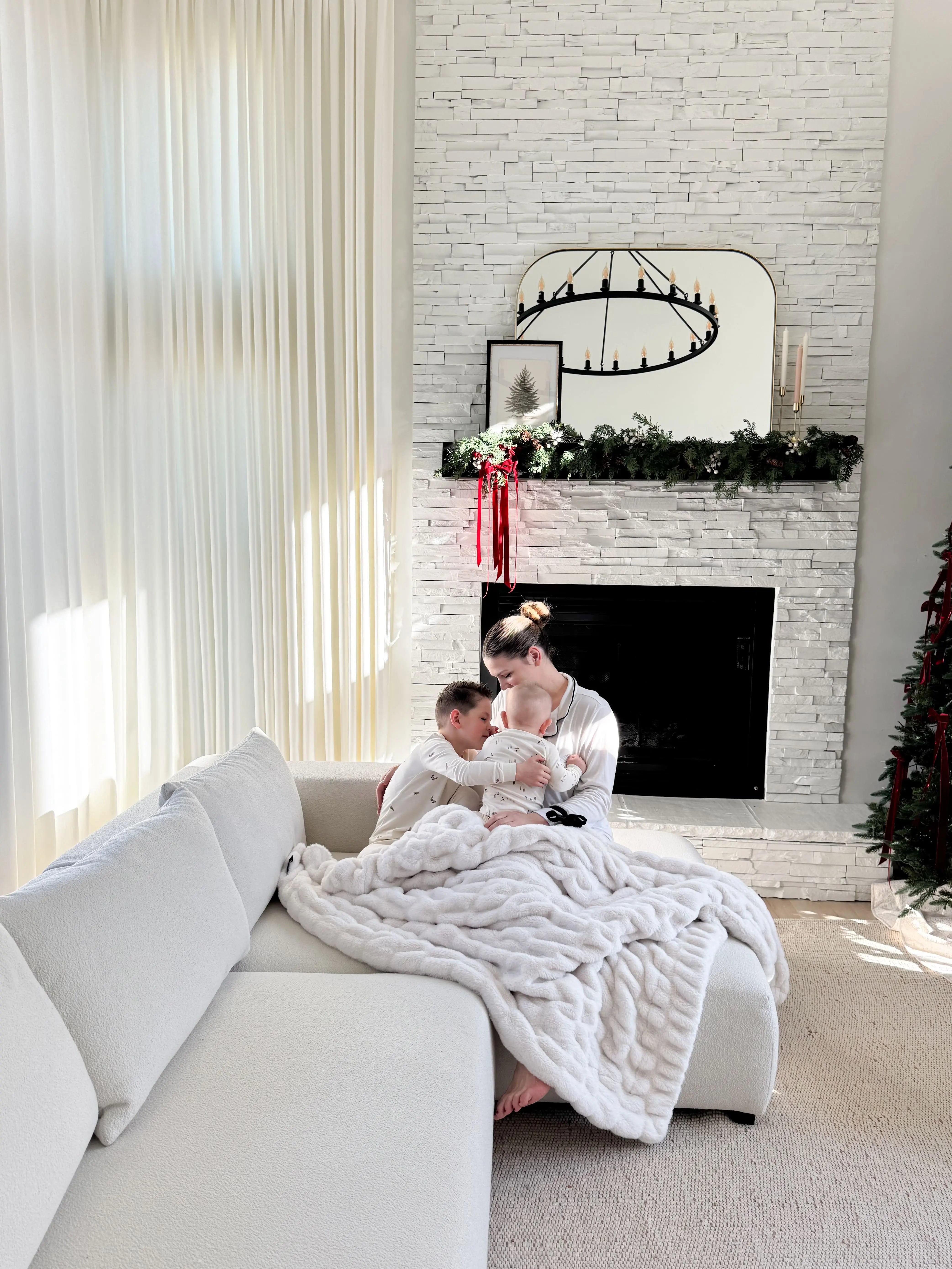 A cozy living room scene with a woman and child sitting on a white sectional sofa wrapped in a chunky white knit blanket, with a white brick fireplace and holiday decor in the background | Influencers Welcome